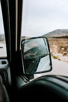 Close-up of car rearview mirror with rain drops reflecting a road. Outdoor travel scene.