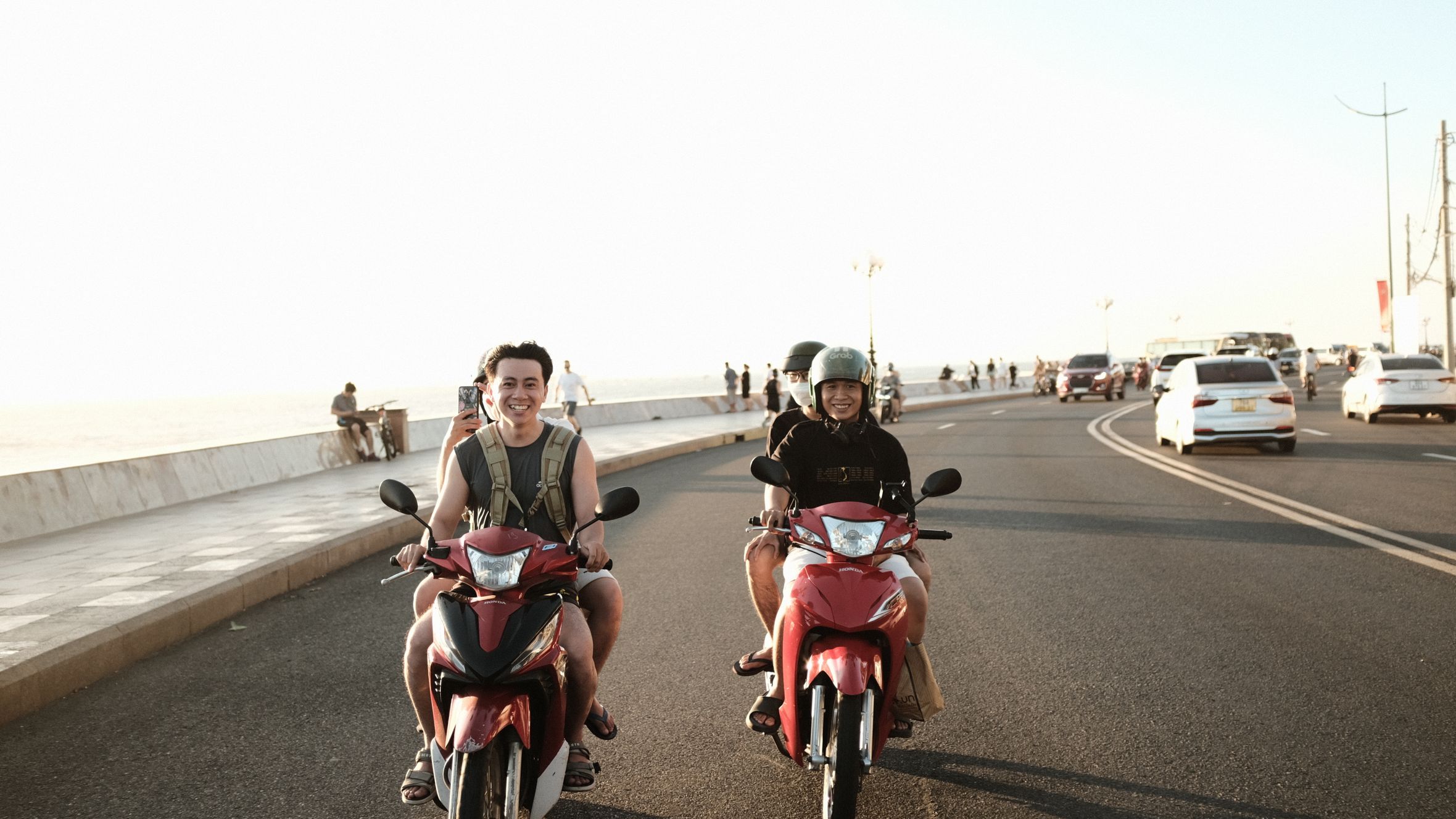 Two young men riding scooters on a sunny Vung Tau coastal road, enjoying the summer day.