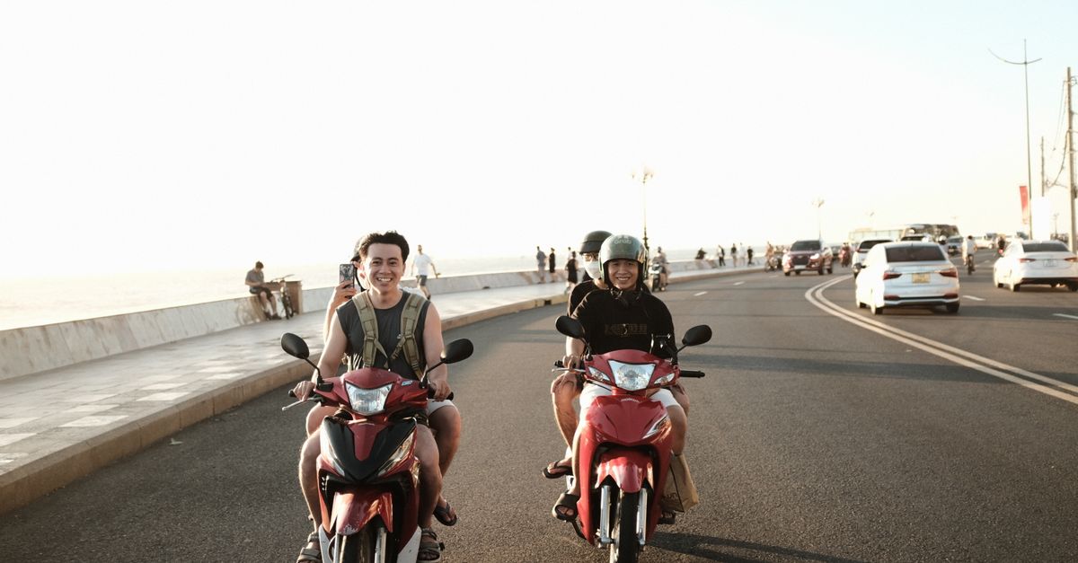 Two young men riding scooters on a sunny Vung Tau coastal road, enjoying the summer day.