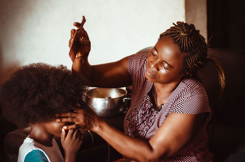 A Black mother parts and combs her child's afro at home, working close to the scalp.