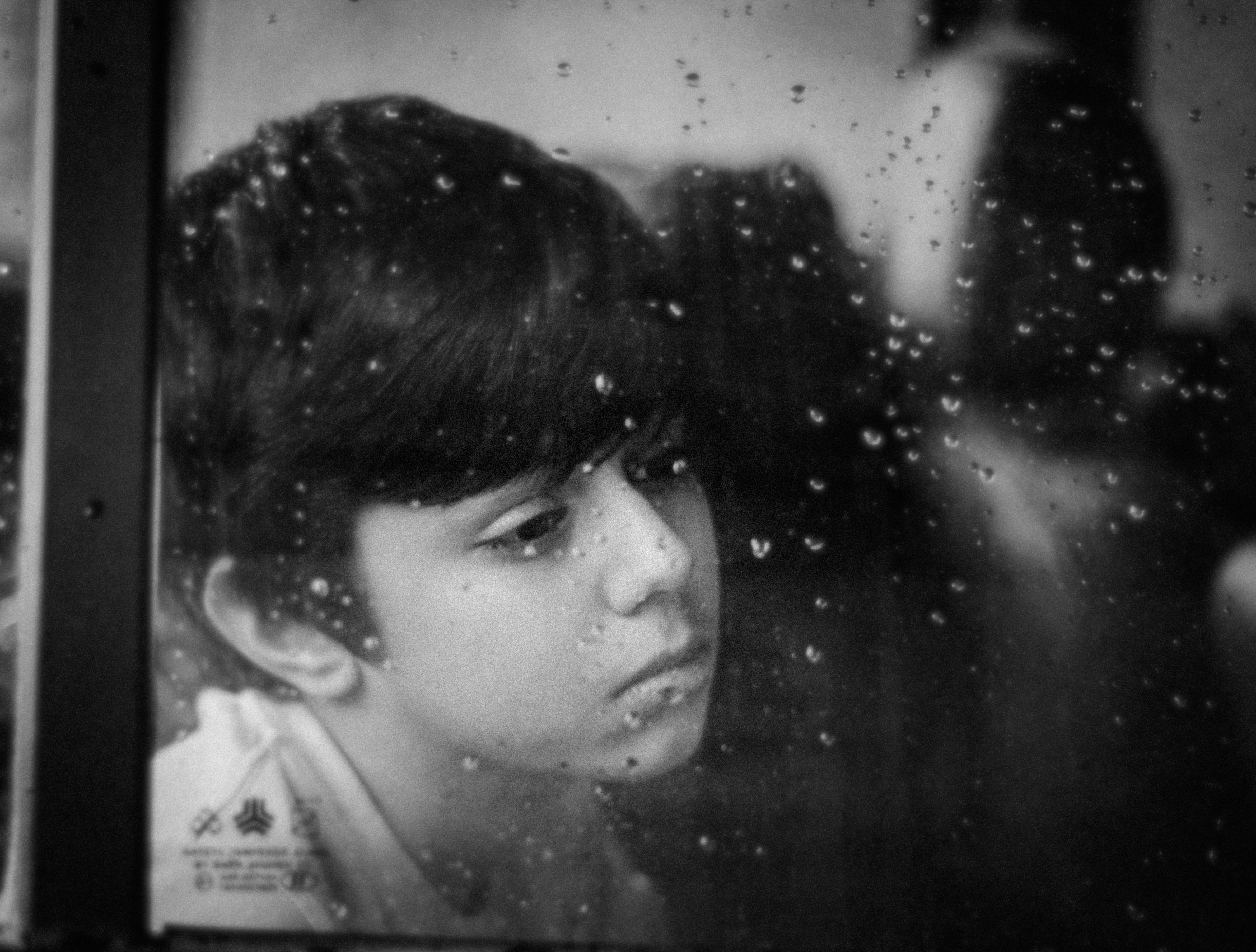 Black and white headshot of a child looking through rain-covered window with a thoughtful expression.