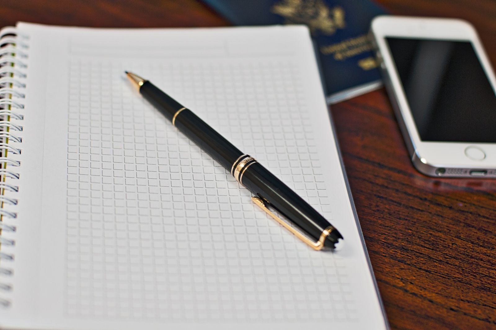 An open spiral notebook with blank grid pages and a black pen resting on it, beside a passport and a smartphone on a wooden table
