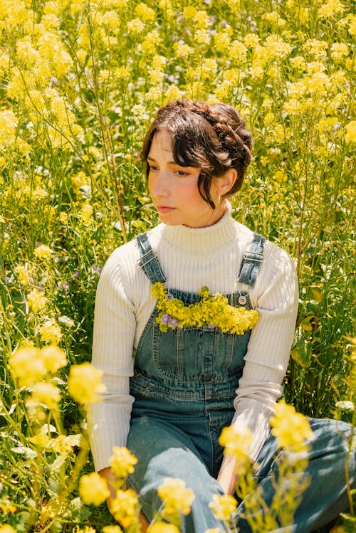 黄色い花畑に座っているデニム ダンガリーの若い女性