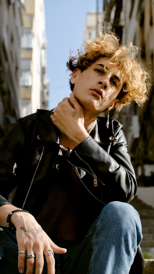 Free Portrait of a fashionable young man with curly hair in a leather jacket sitting outdoors. Stock Photo