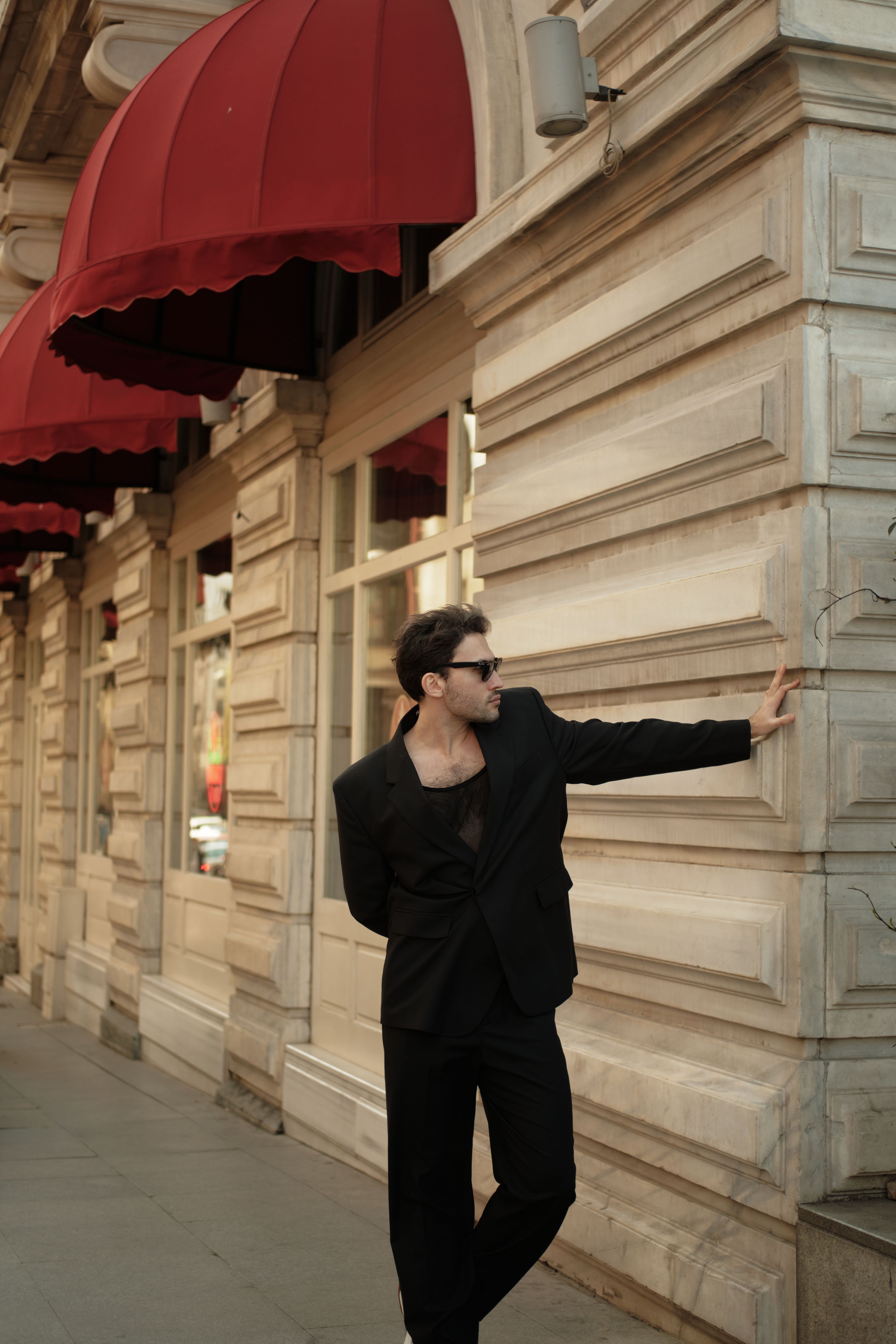 Man in fashionable attire poses against a tenement building with red awnings, showcasing urban style.
