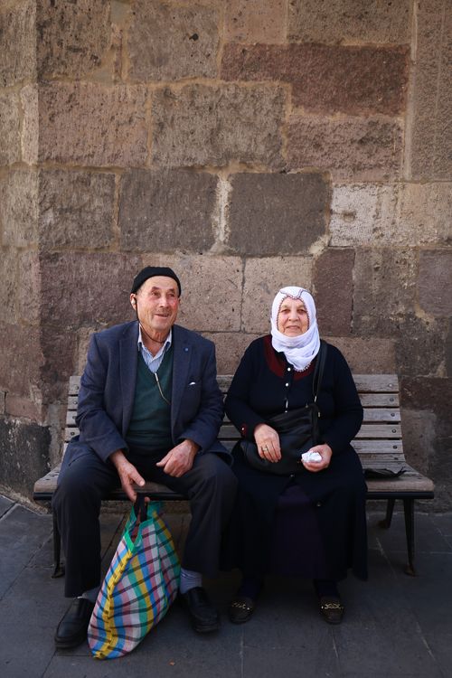 Free Senior couple sitting on a bench against a stone wall, enjoying a sunny day. Stock Photo