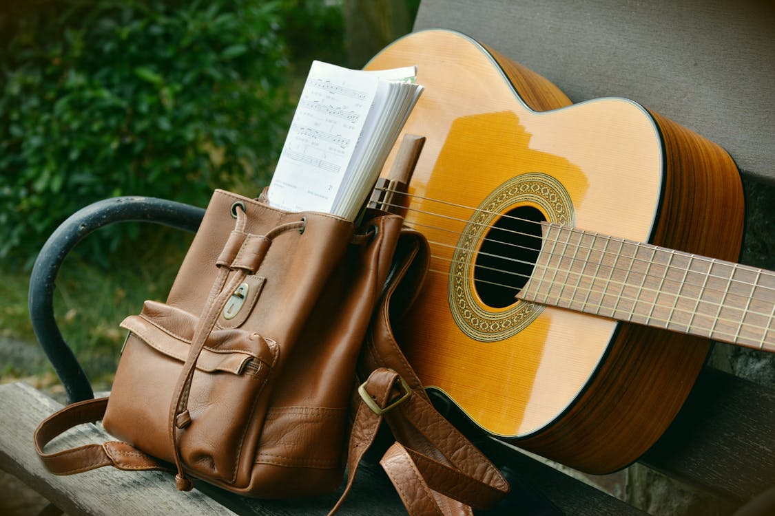 Free Brown Acoustic Guitar Beside Brown Leather Bucket Backpack on Brown Wooden Bench Stock Photo