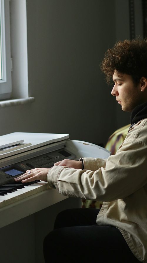 Free A young man plays an electronic keyboard by a window, focused and intent. Stock Photo
