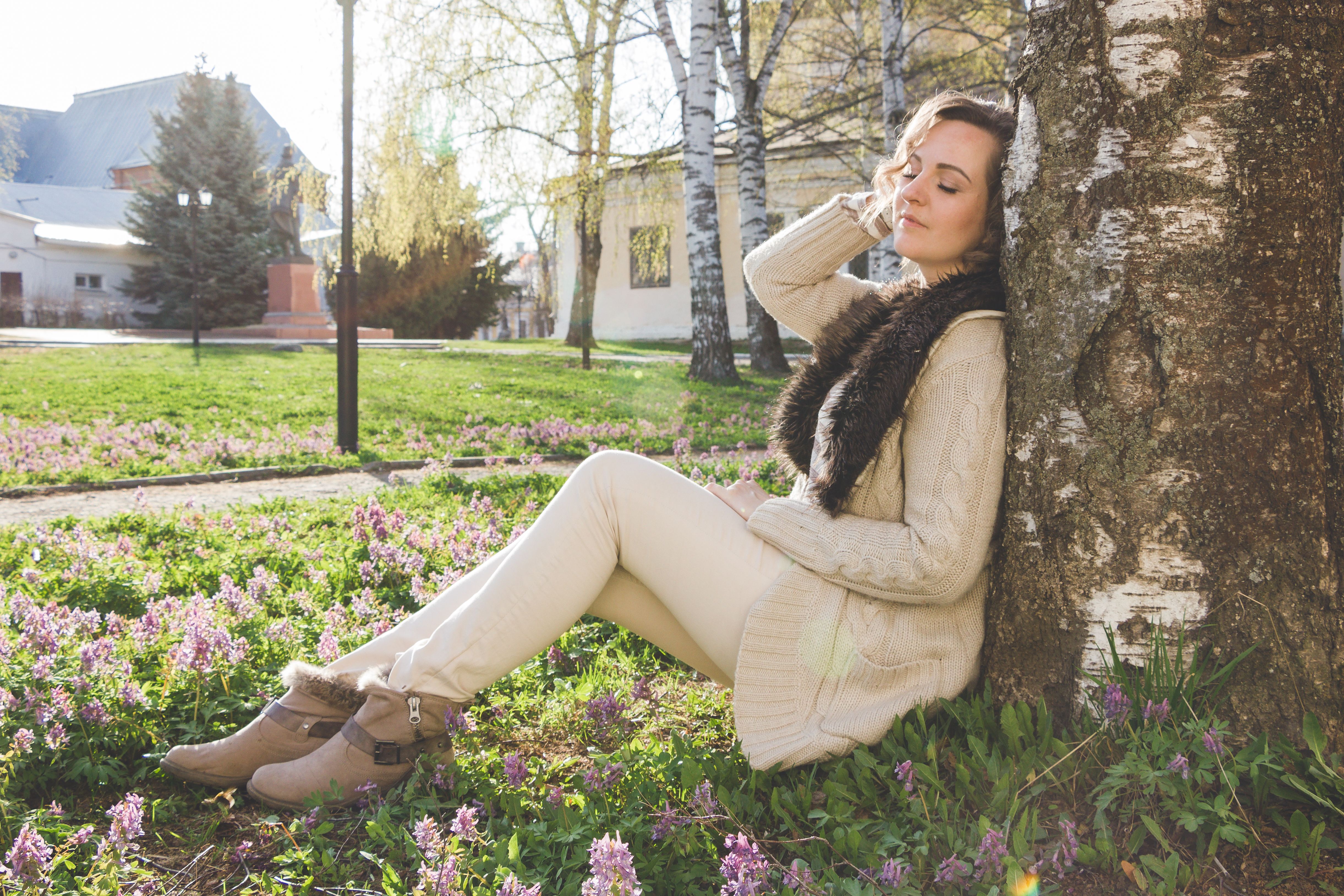 A woman enjoys a peaceful moment sitting against a tree in a spring park, surrounded by blooming flowers.