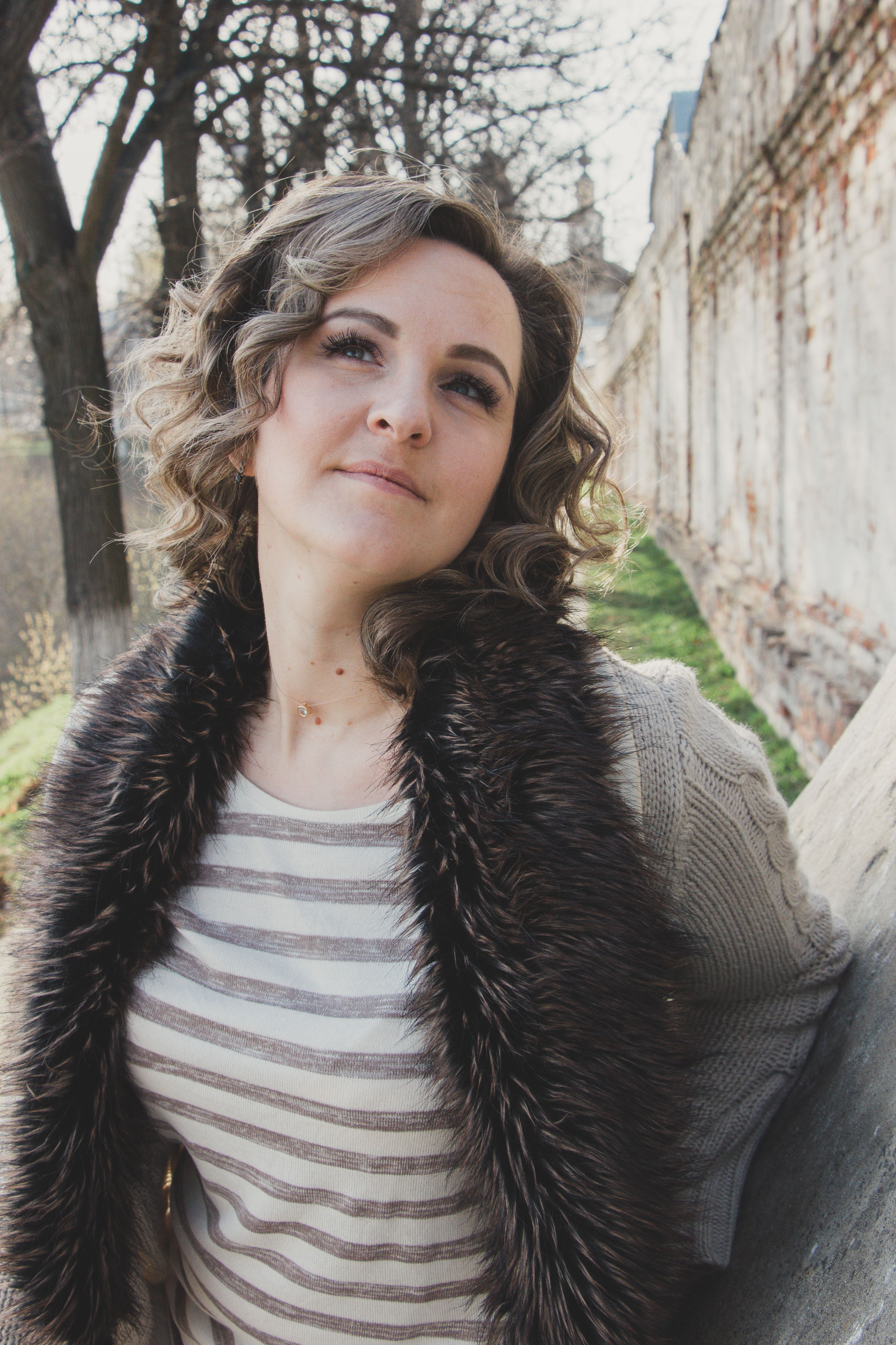 Stylish woman posing near an old brick wall, embracing the cool weather with her cozy fur-lined sweater.