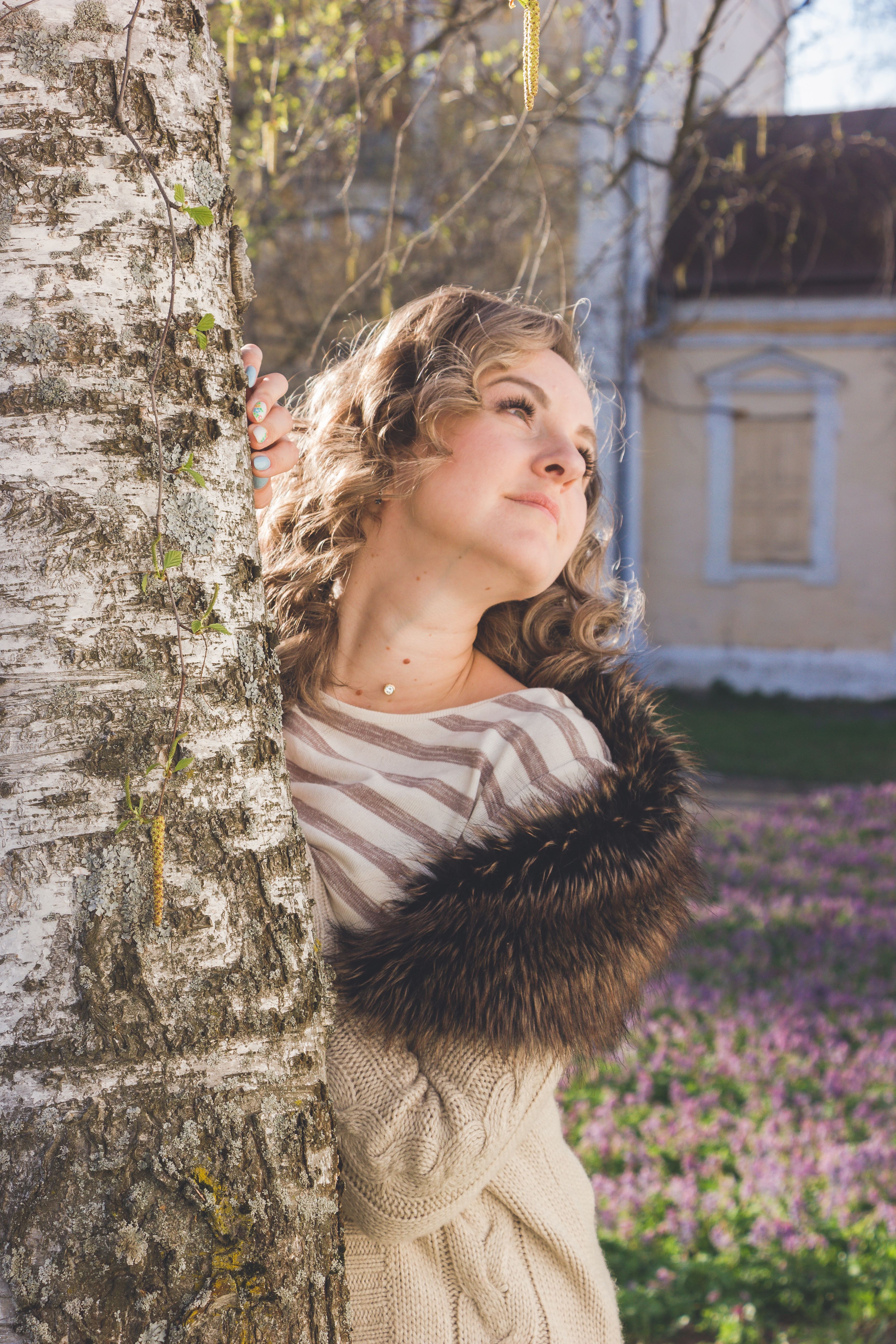 Woman in a fur-trimmed cardigan leaning against a tree with a serene expression during springtime.