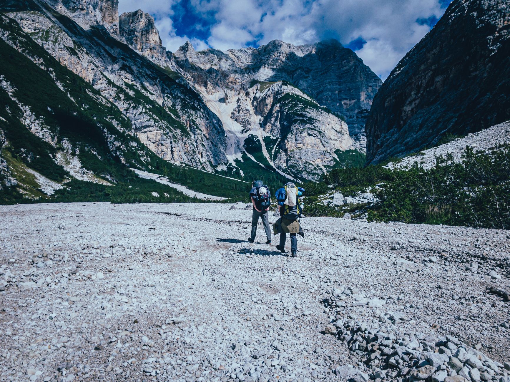 Two hikers navigate a rocky trail surrounded by breathtaking mountain scenery in daylight.
