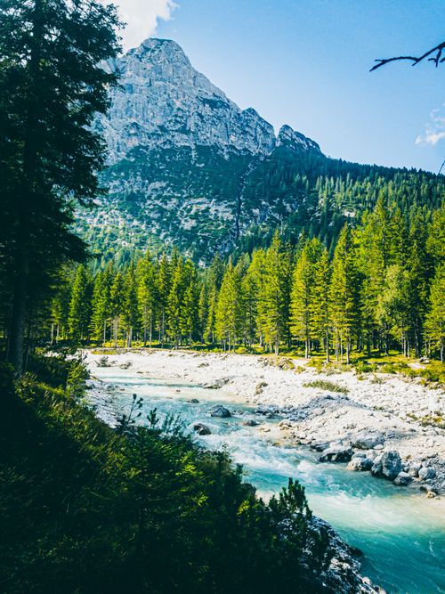 Free Beautiful alpine landscape featuring a mountain, lush forests, and a serene river under clear skies. Stock Photo