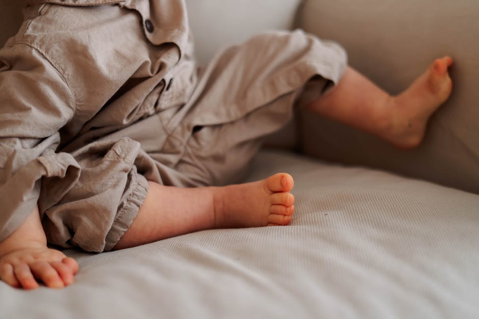 A cozy close-up of a baby in neutral gray clothes sitting barefoot on a sofa.