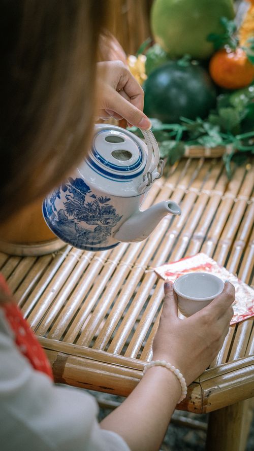 Free A woman pouring tea from a blue ceramic teapot into a cup on a bamboo table. Stock Photo