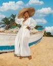 穿着白色连衣裙、戴着帽子的时尚女人站在一艘海滩船附近
