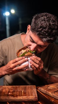 A man indulges in a large burger outdoors under night lights, wrapped in paper.
