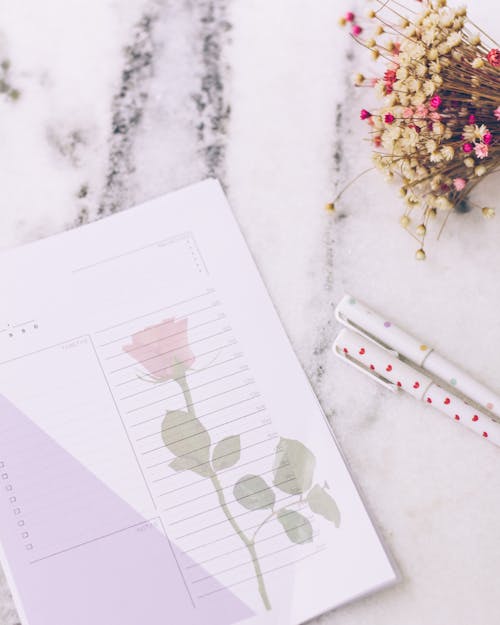 A Piece of Paper with an Illustration of a Rose, Pens and Flowers Lying on a Marble Counter 