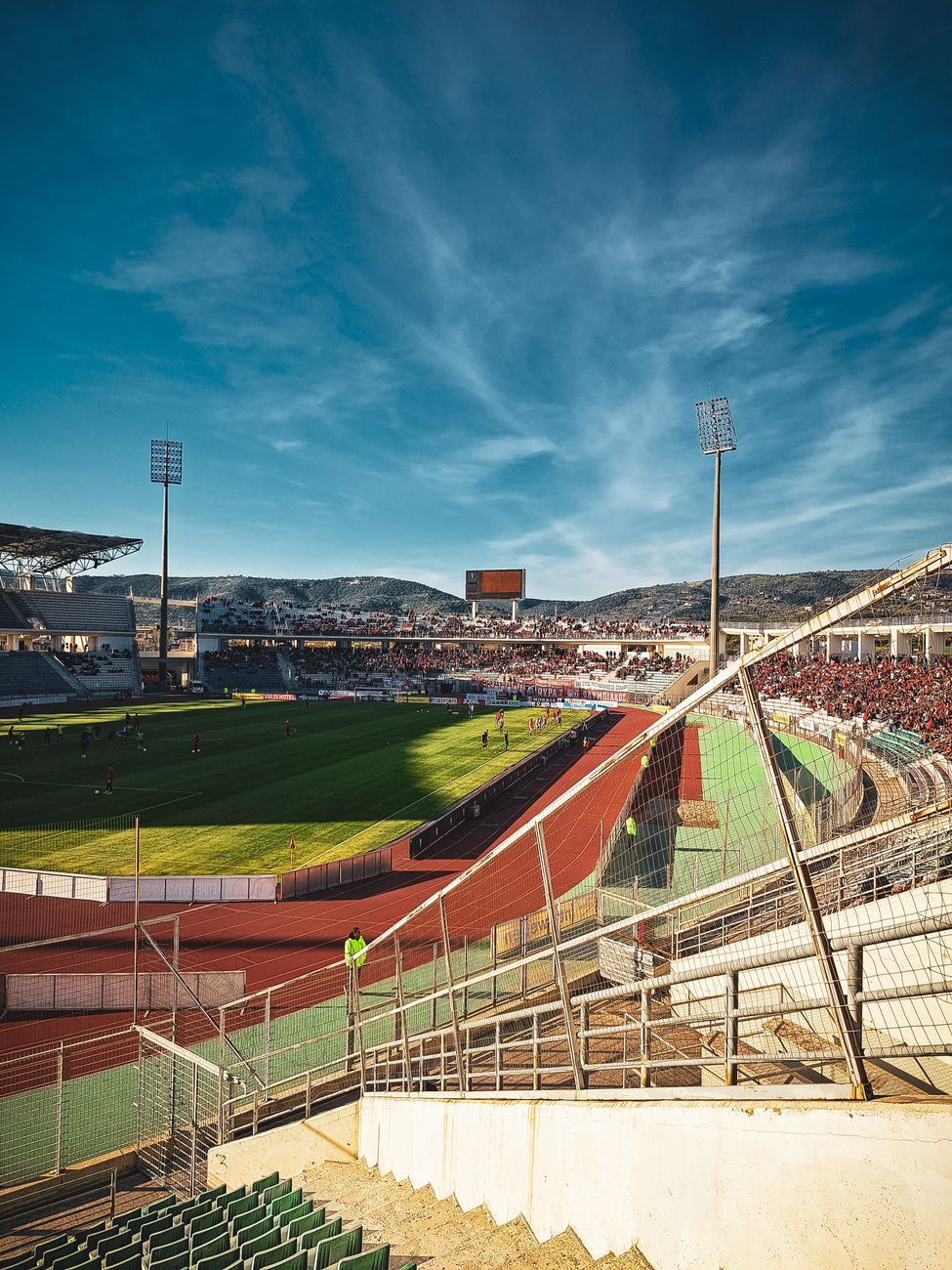 Een levendig tafereel in het Panthessaliko Stadion in Volos, Griekenland, met toeschouwers die genieten van een zonnige dag.