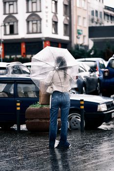 A woman stands in a rainy city street holding a transparent umbrella.