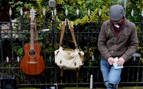 白と茶色のスリングバッグと茶色のアコースティックギターの横にある黒い鋼のフェンスに寄りかかって男