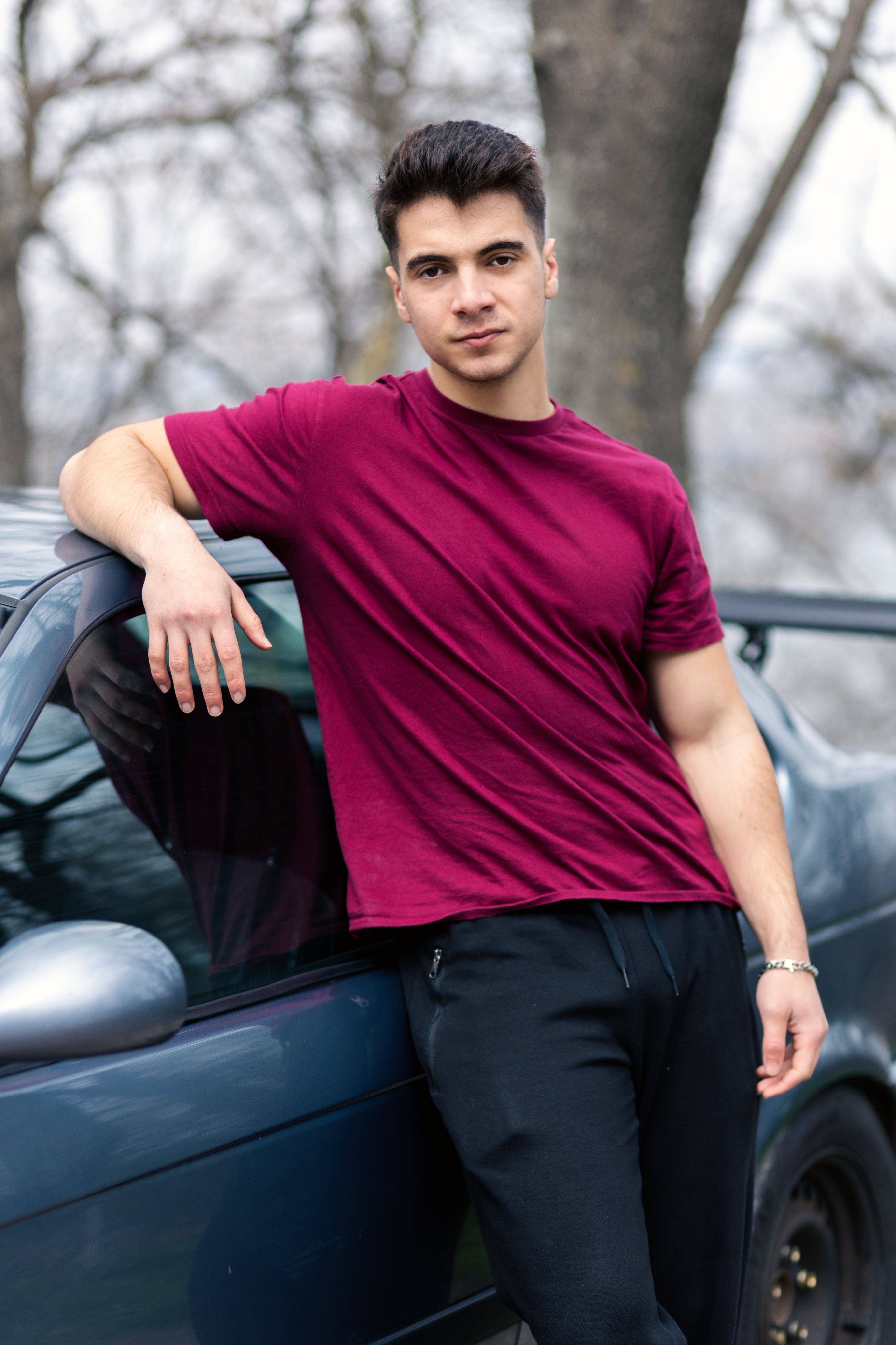 A young man in a burgundy t-shirt leans casually against a car outdoors.