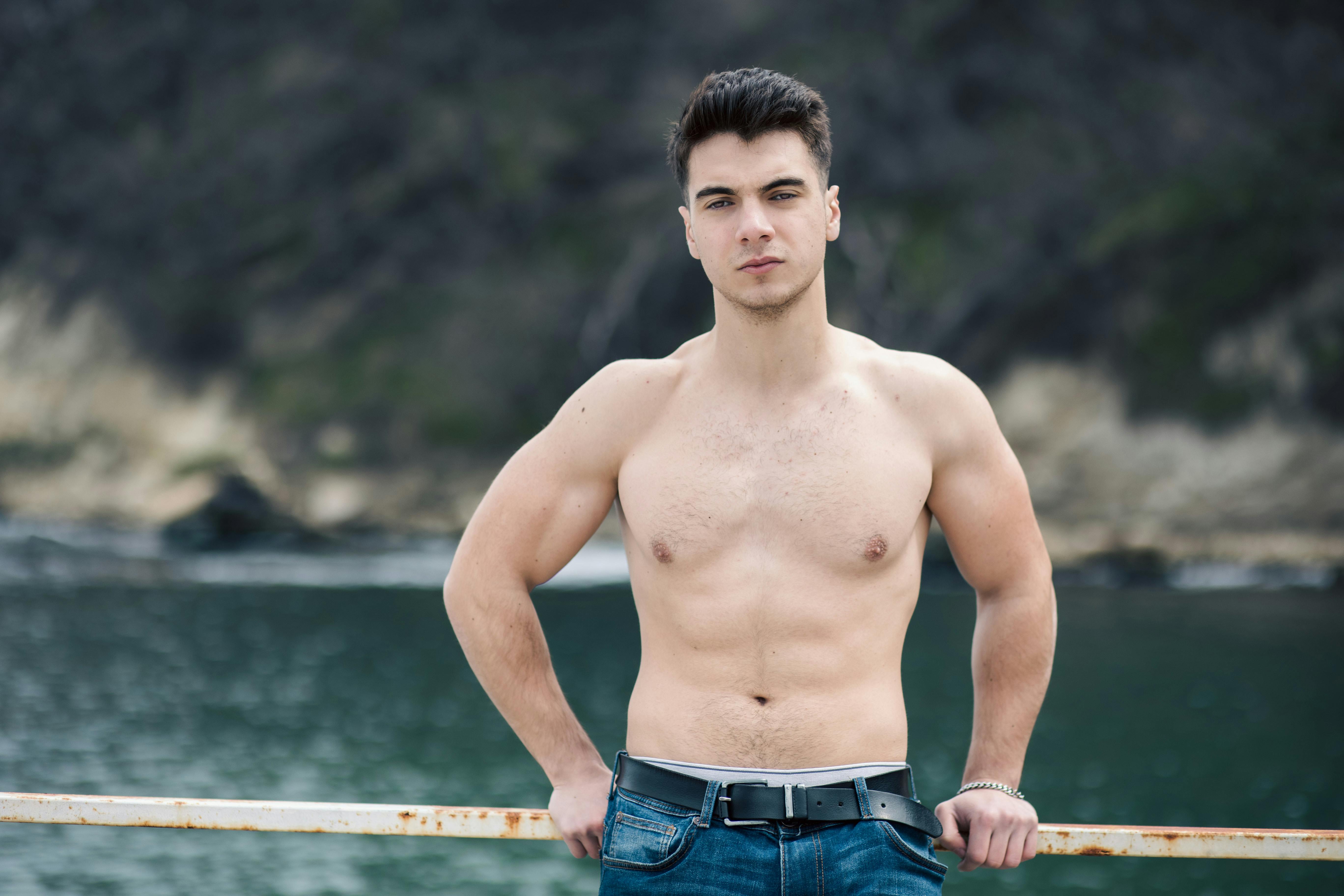 Athletic young man posing shirtless by the waterfront, showcasing physique.