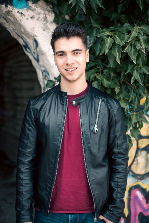 Free Young man wearing a leather jacket and t-shirt smiling outdoors by ivy-covered wall. Stock Photo