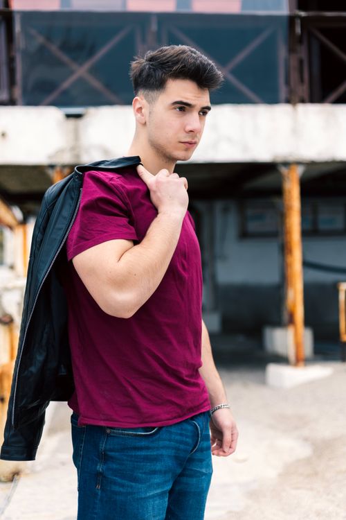 Free Young man in casual attire posing confidently outdoors with leather jacket draped over shoulder. Stock Photo