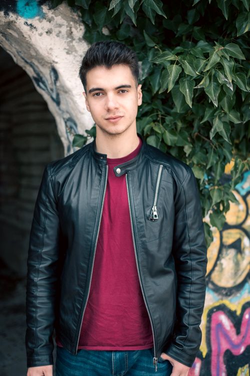 Free Portrait of a fashionable young man in a leather jacket standing next to a graffiti wall. Stock Photo
