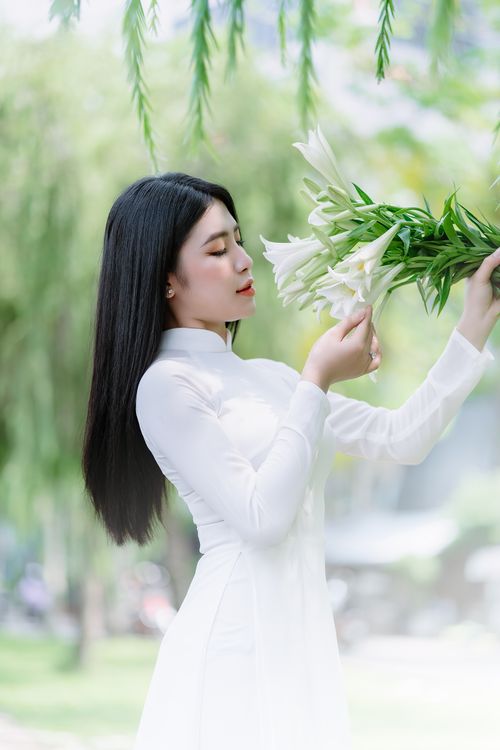 Free Asian woman in white dress holding lilies, posing elegantly outdoors in a serene summer setting. Stock Photo