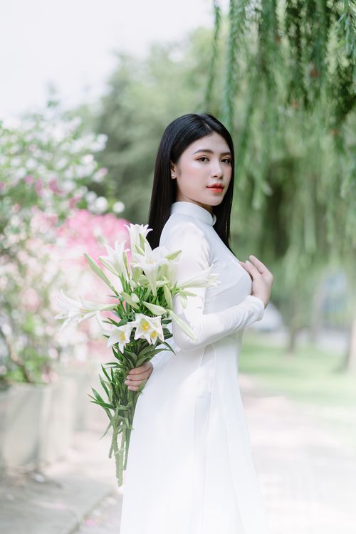 Free Young Asian woman in white dress posing with bouquet of white lilies outdoors. Stock Photo