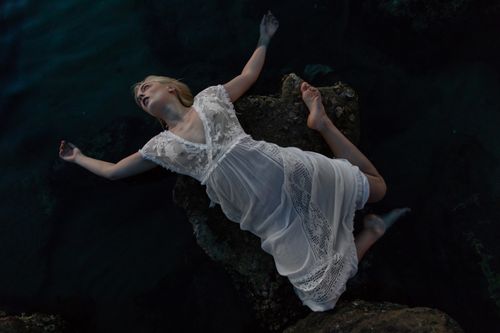 Free A woman in a white lace dress lays on rocks, captured with an artistic, moody tone. Stock Photo