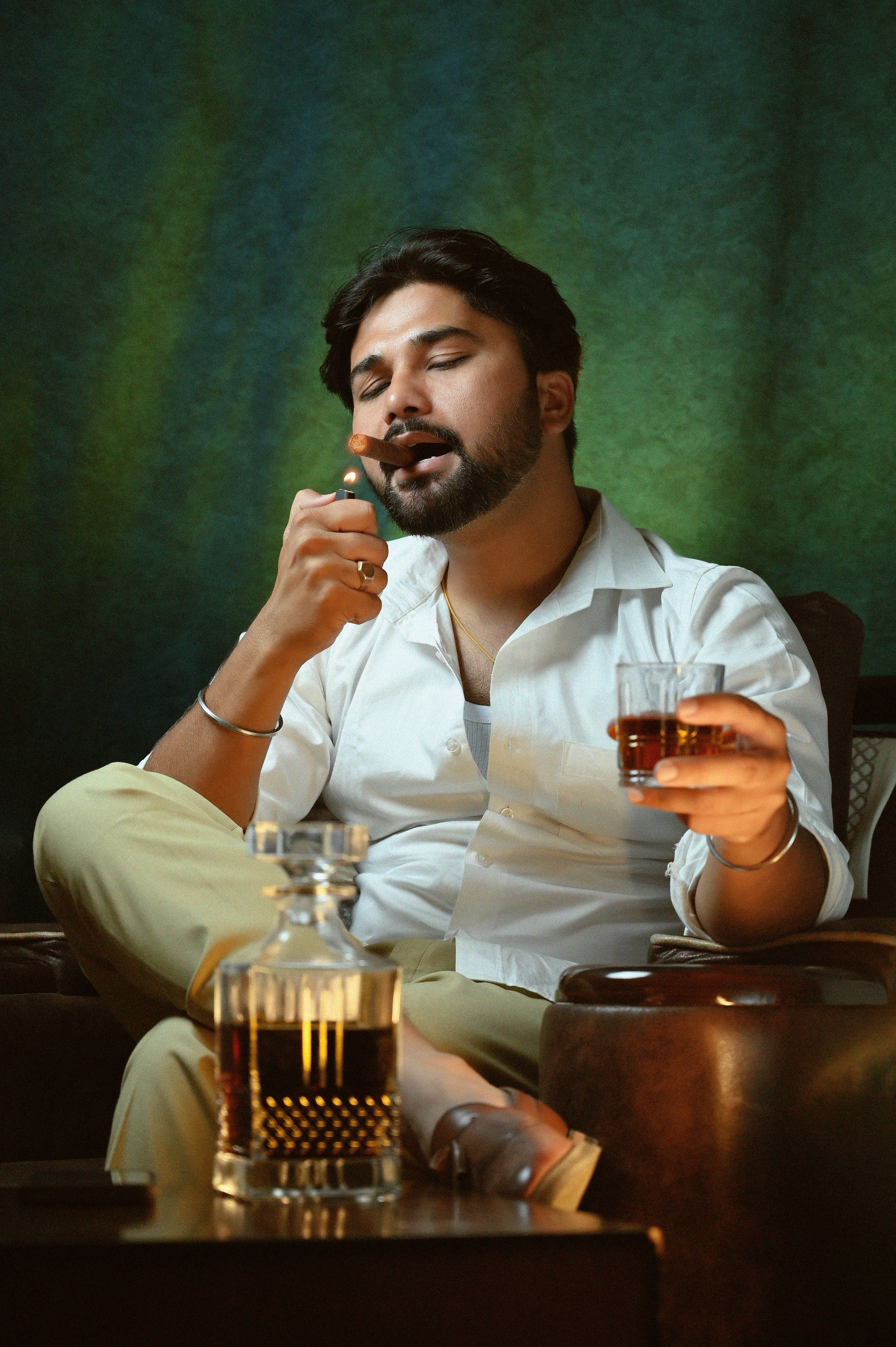 A fashionable man sits indoors enjoying a glass of whiskey and a cigar, exuding style and relaxation.
