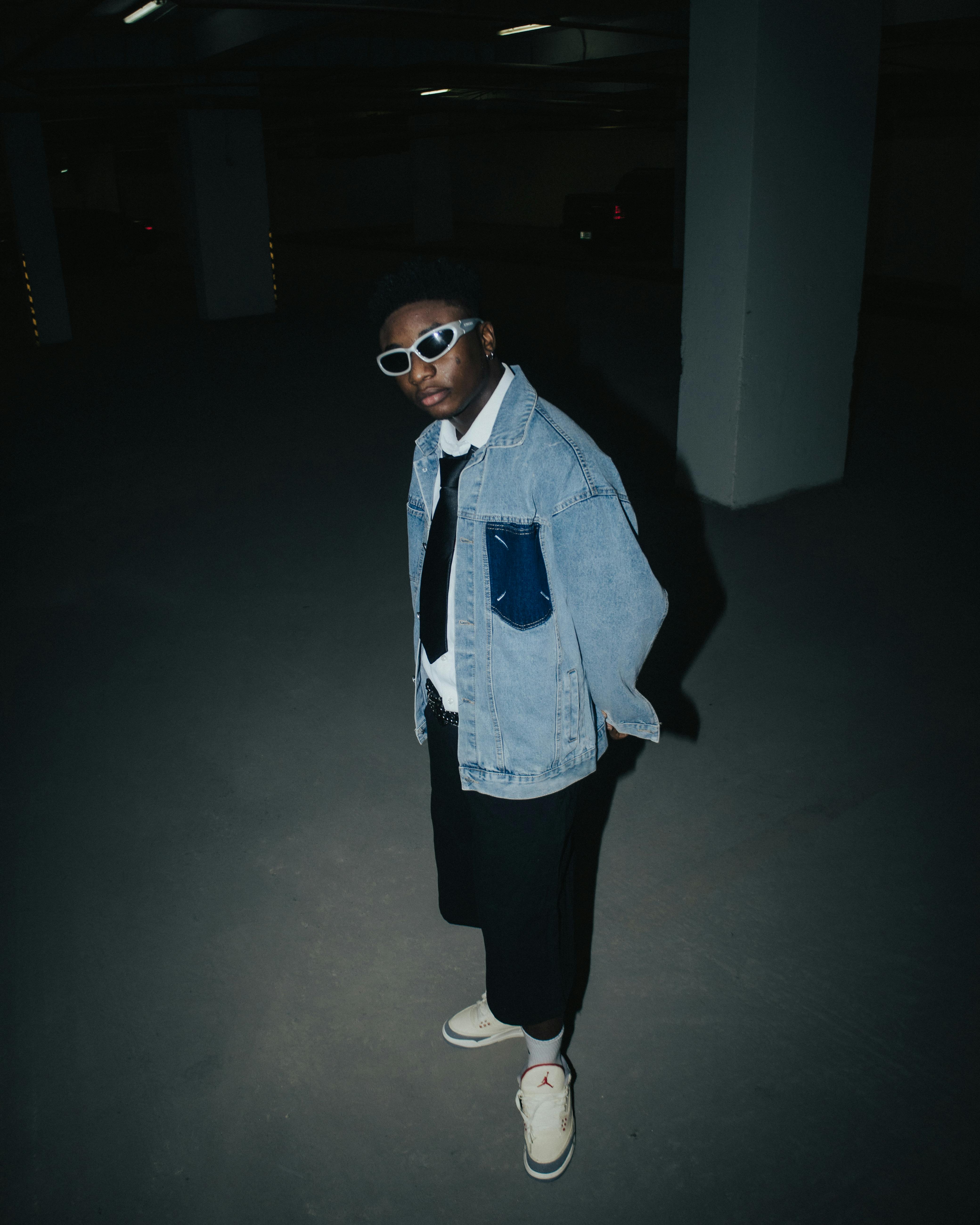 A teenager poses in a denim jacket and sneakers in a dimly lit parking garage.