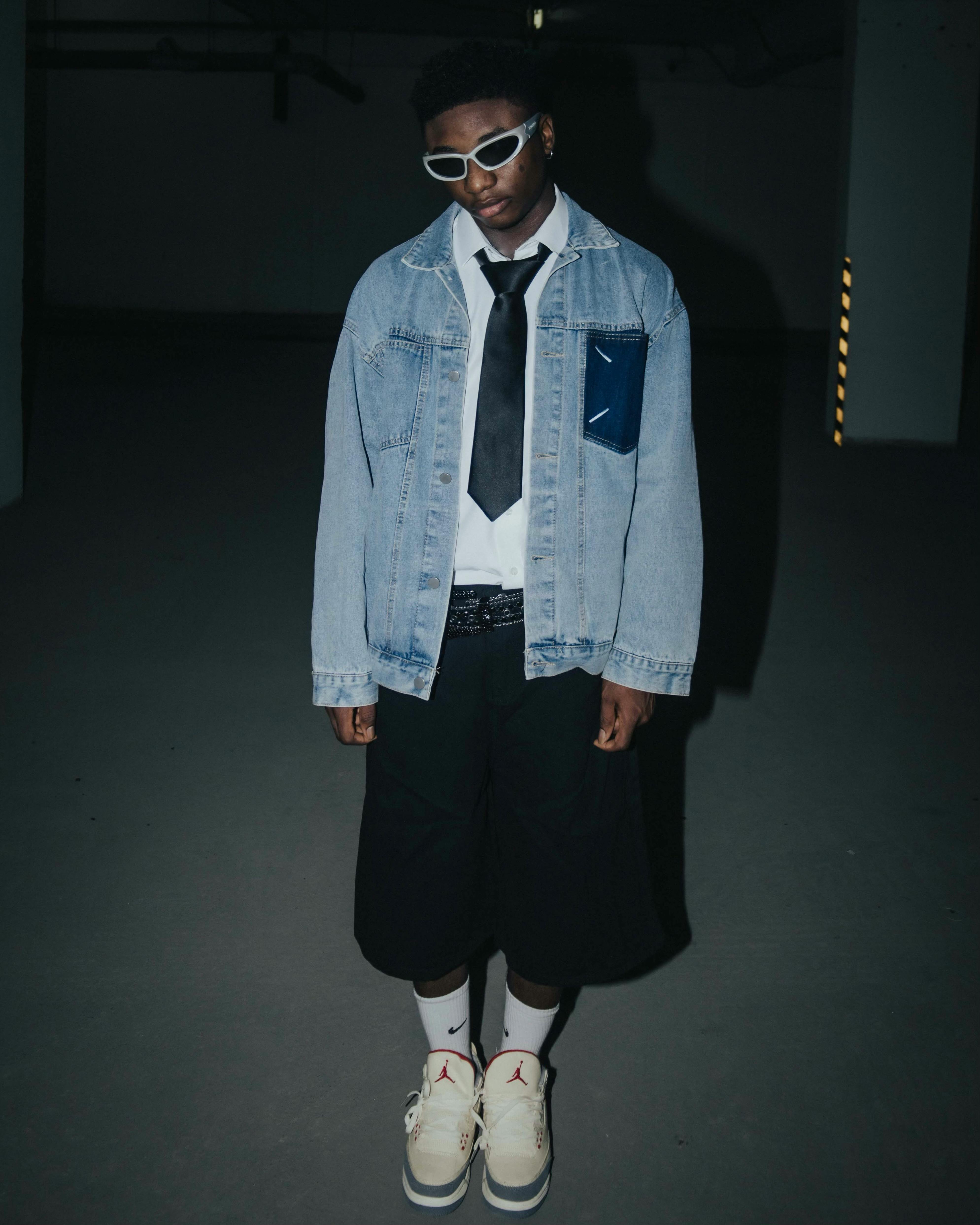 A stylish teenager poses in a denim jacket and sunglasses, exuding confidence in an urban setting at night.