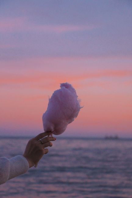 A hand holding pink cotton candy against a pastel sunset over the ocean.