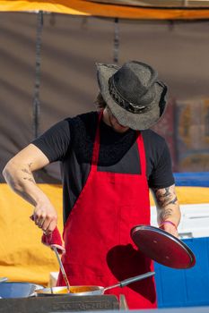 A man in a red apron and cowboy hat cooking at an outdoor festival, adding flavor and excitement.