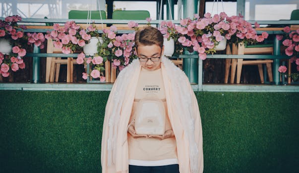 一名戴着眼镜和围巾、背着背包的年轻人站在室内，周围悬挂着粉红色的花朵