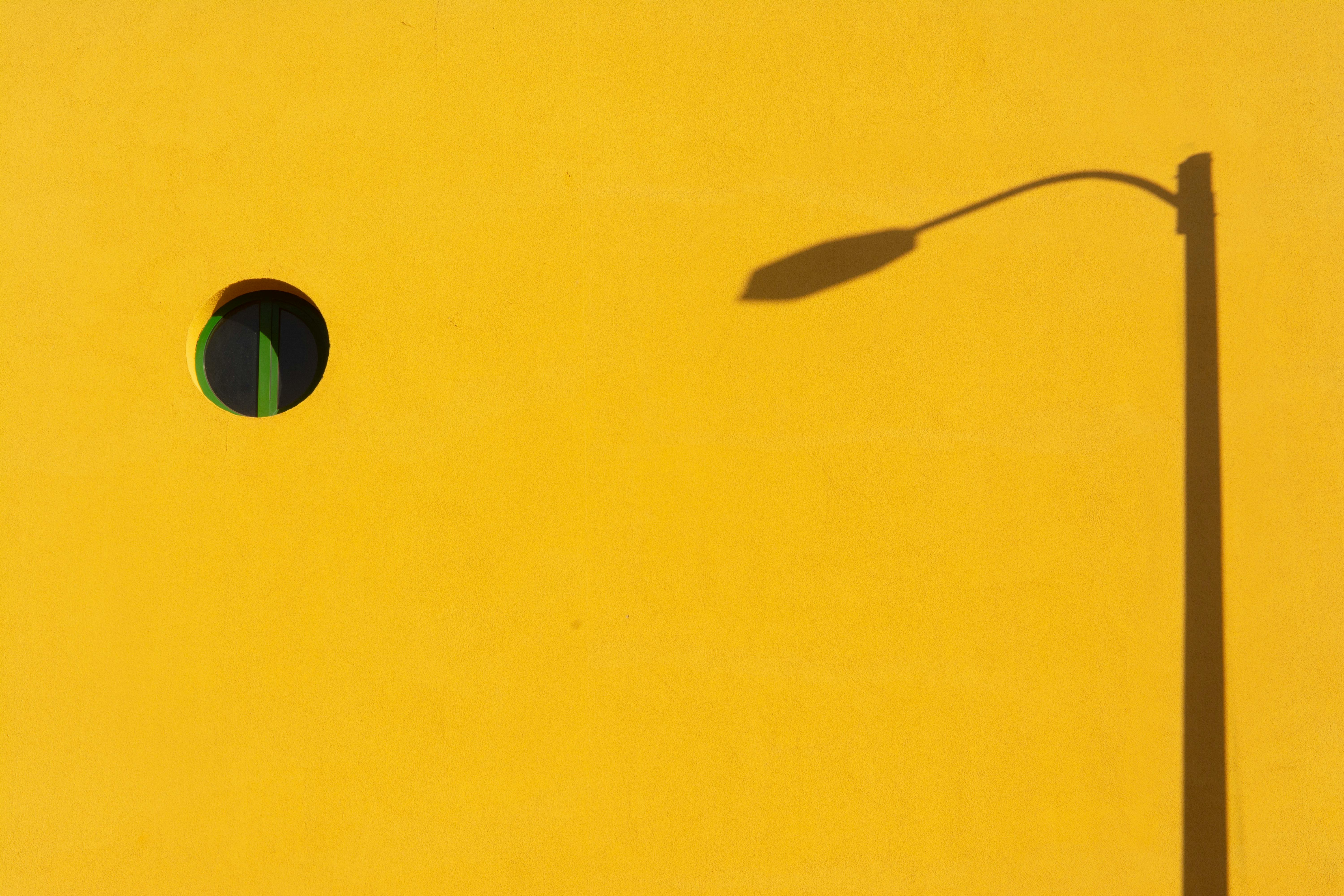 Abstract photo of a yellow wall with a lamppost shadow and round window.