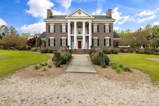 Elegant colonial mansion with columns and a red door, surrounded by lush greenery under a blue sky.