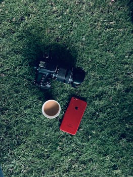 Flat lay of a camera, coffee cup, and red smartphone on grass.