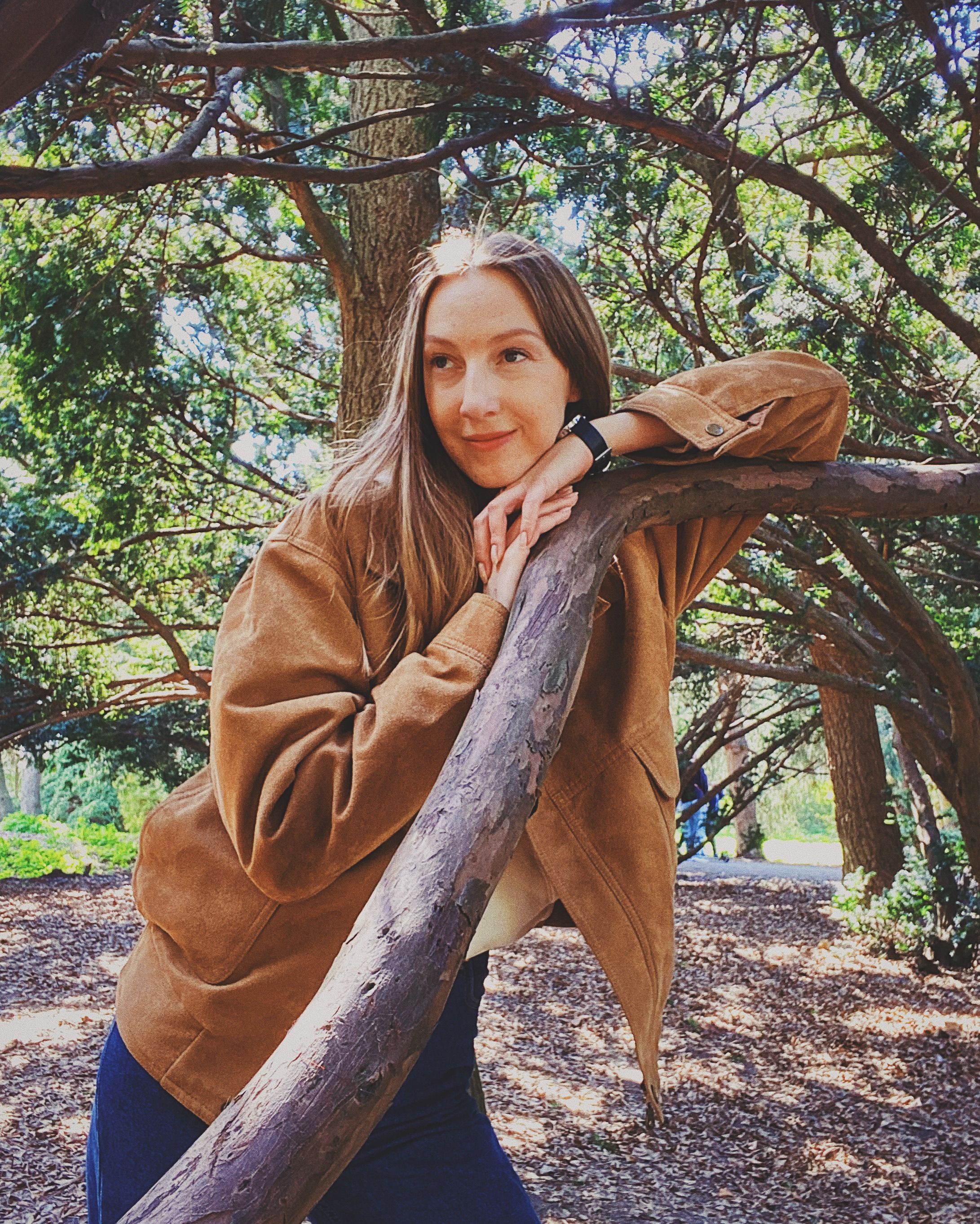 Woman with long hair leans on tree branch in sunny Kyiv park, wearing casual jacket.