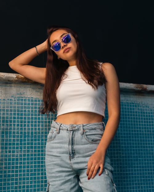 Free Young woman in white crop top and jeans, leaning against mosaic wall, sunglasses on. Summer fashion. Stock Photo