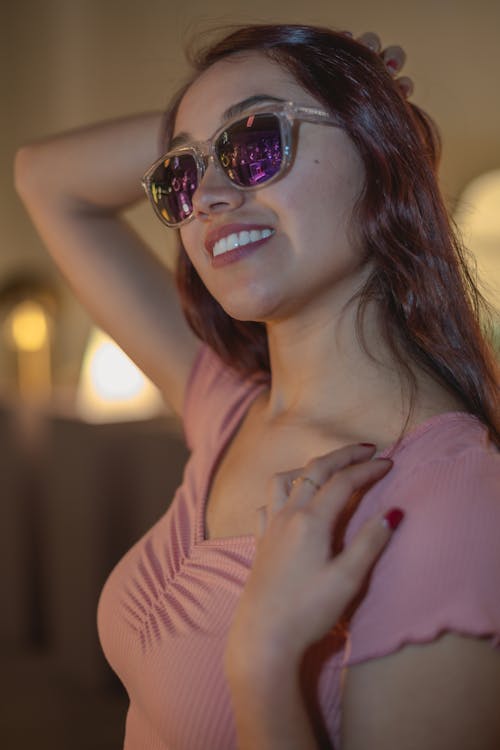 Young Woman in a Pink T-shirt and Sunglasses Standing with Hand in Hair and Smiling 