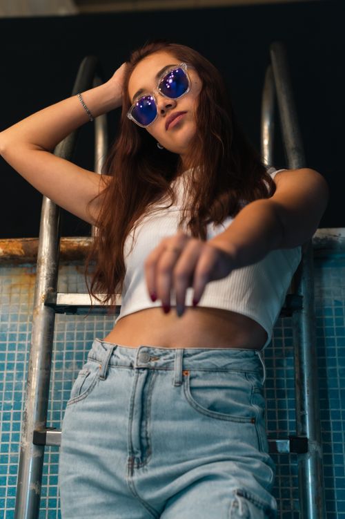 Free A fashionable woman in sunglasses and a crop top posing confidently by a swimming pool ladder. Stock Photo