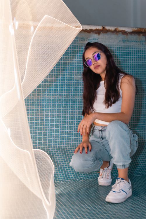 Free Young woman wearing sunglasses poses stylishly against a blue-tiled wall in fashion photography. Stock Photo