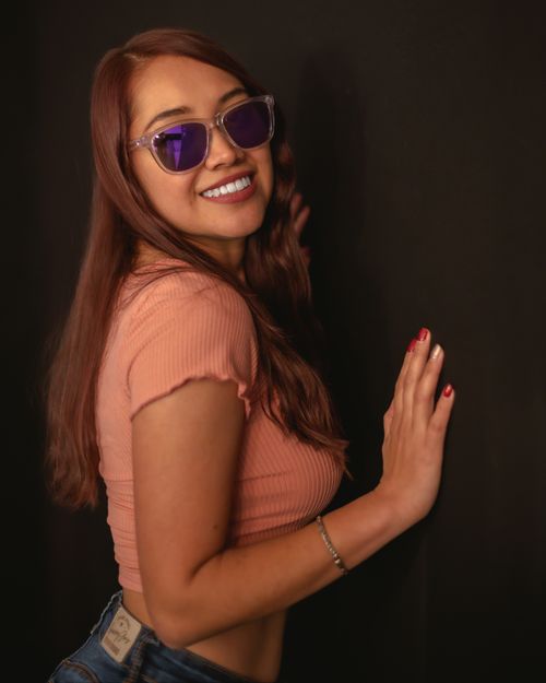 Free Red-haired woman wearing purple sunglasses and a pink crop top, smiling and posing against a dark background. Stock Photo