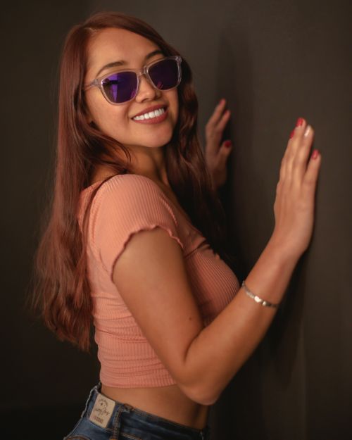 Free Young woman in a trendy crop top and sunglasses posing confidently indoors with a vibrant smile. Stock Photo
