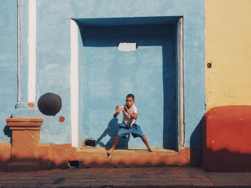 Free A child joyfully playing with a ball against a colorful blue wall in a sunny outdoor setting. Stock Photo